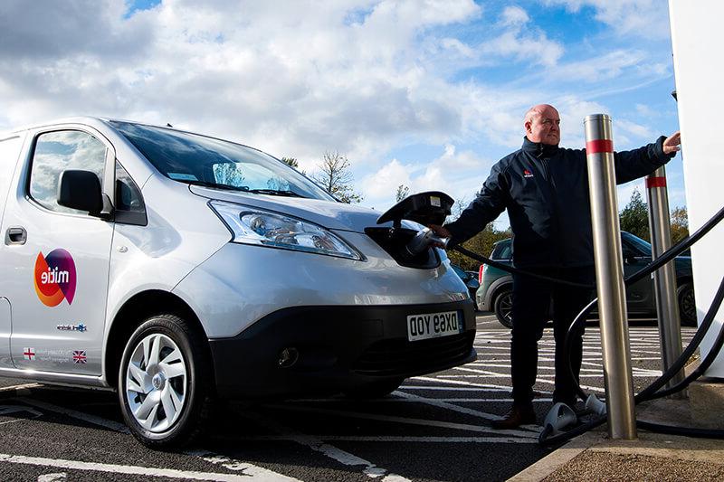 一辆米蒂牌电动货车正在充电, with a Mitie employee operating the charge point system at the front of the van